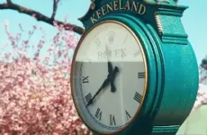 Teal clock tower at Keeneland racecourse with Roman numerals, pink blossoms in the foreground and a blue sky backdrop.